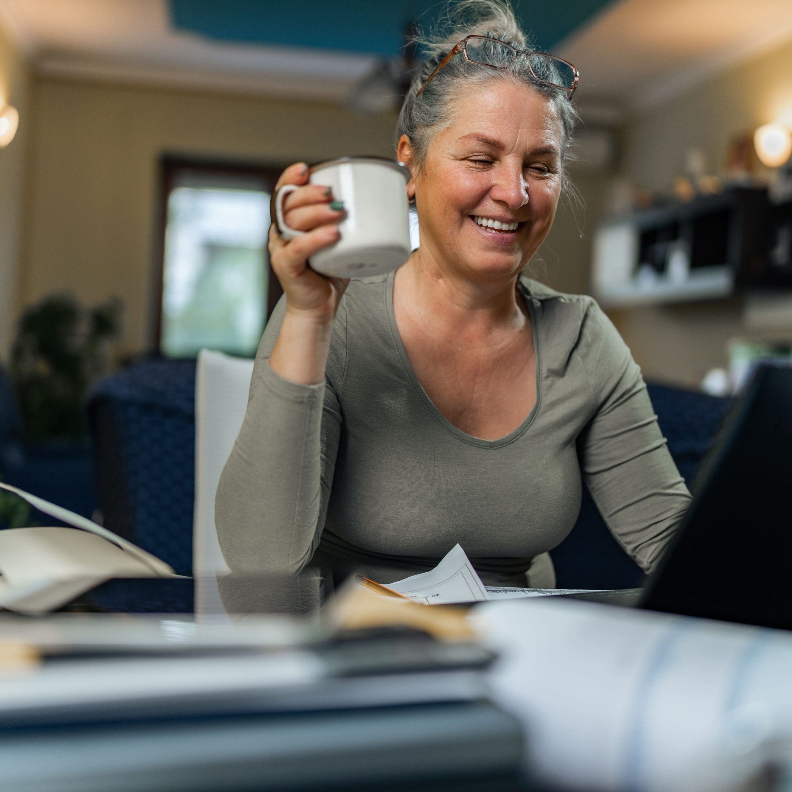 casually dressed senior woman with gray hair sitting at working table taking to friend via electronic device , drinking coffee