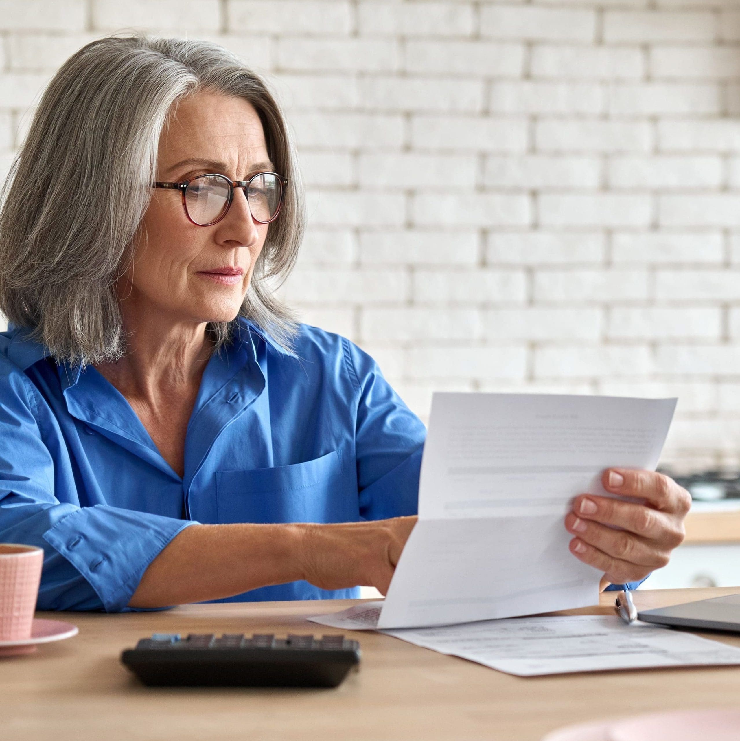 Adult senior 60s woman working at home at laptop. Serious middle aged woman at table holding document calculating bank loan payments, taxes, fees, retirement finances online with computer technologies
